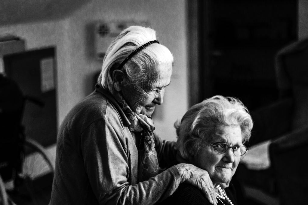 Portrait of two older ladies, one standing with hand on the shoulder of the one sitting down